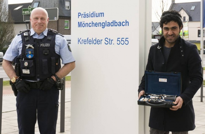 Zwei Männer stehen recht und links von einer Säule mit der Aufschrift Präsidium Mönchengladbach. Einer der beiden trägt eine Polizeiuniform. Der andere hält eine Klarinette in der Hand.