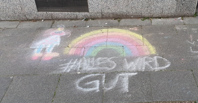 Ein Regenbogen, mit Kreide auf die Straße gemalt und zum Teil verwischt. Darunter steht in Großbuchstaben: Alles wird gut.