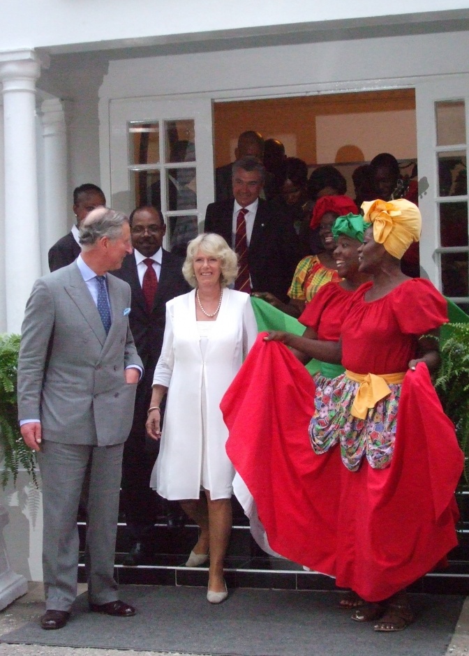 Prinz Charles und Herzogin Camilla stehen nebeneinander in einem Hauseingang, neben ihnen 2 dunkelhäutige Frauen in traditionellen jamaikanischen Kleidern