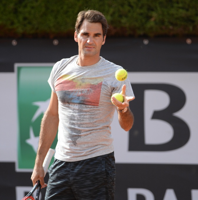 Roger Federer in T-Shirt und Shorts auf dem Tennisplatz. Er hält einen Tennisschläger in der einen und einen Tennisball in der anderen Hand.