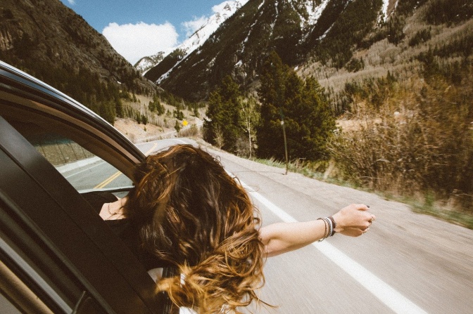 Ein Auto fährt über eine Straße. Eine Frau hängt sich aus dem Autofenster  und lässt ihre Haare im Wind wehen.