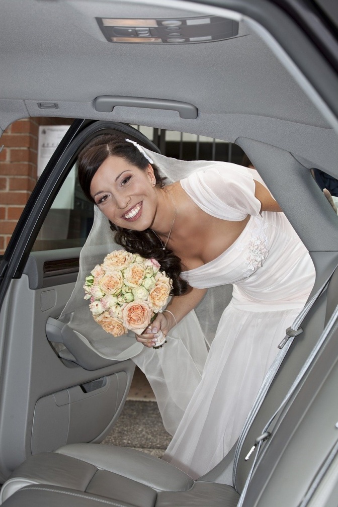 Eine Braut in Hochzeitskleid und mit Brautstrauß streckt ihren Kopf durch eine geöffnete Autotür.