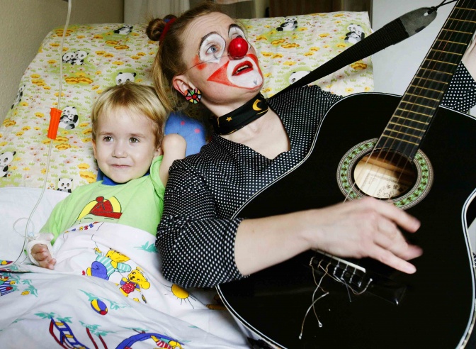 Eine Clownin liegt mit einem Kind in einem Krankenhausbett und spielt Gitarre.