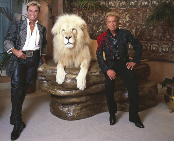 2 Männer stehen rechts und links von einem weißen Löwen auf einem Podest.