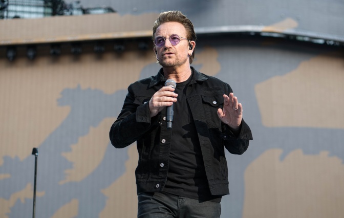 Bono mit getönter Brille auf der Bühne. Er singt in ein Mikrophon.