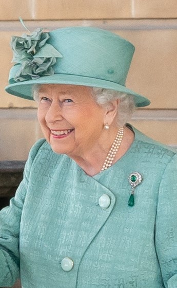 Die Queen mit kurzen, gelockten, weißen Haaren. Hut und Kostüm sind aufeinander abgestimmt.