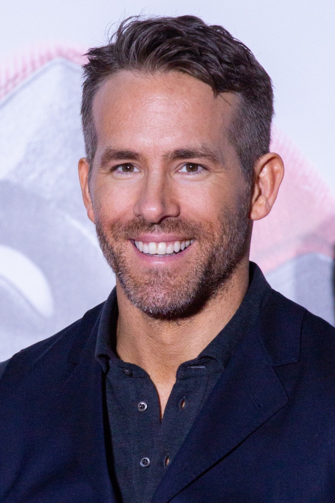 Ryan Reynolds lächelnd vor einer Logowand. Er hat kurze Haare und trägt einen. Dreitagebart.