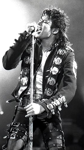 Michael Jackson mit Locken im Bühnenoutfit. Er singt in ein Standmikro.