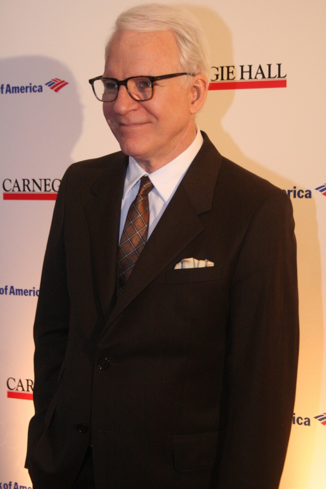 Steve Martin mit weißen Haaren und Brille. Er steht in Anzug und Krawatte vor einer Logowand.
