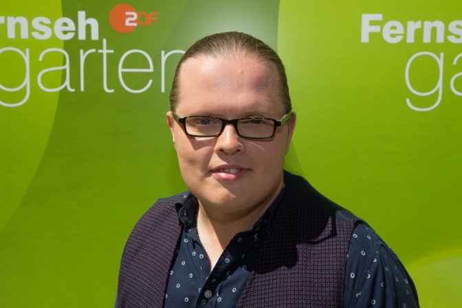 Angelo Kelly mit Brille und zum Zopf gebundenen Haaren vor der Logowand des ZDF Fernsehgartens