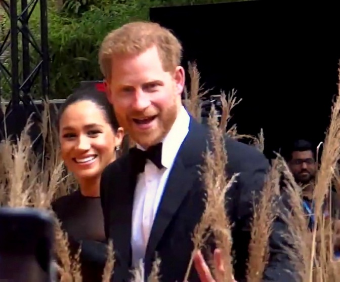 Prinz Harry und Herzogin Meghan in festlicher Kleidung. Sie lächeln und laufen an Schilfgras vorbei.