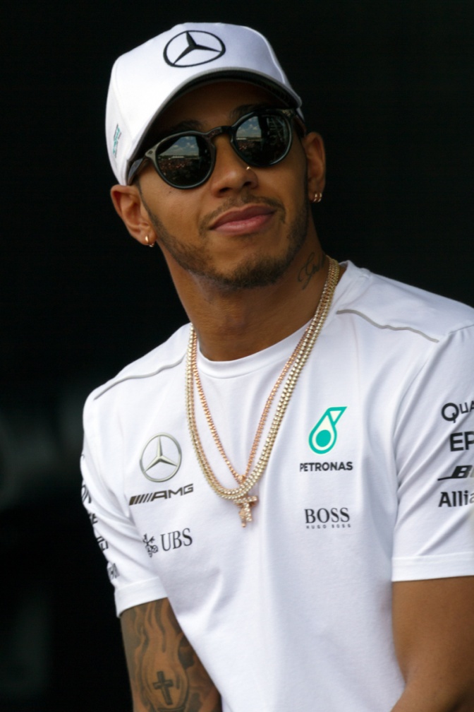 Lewis Hamilton mit T-Shirt und Schirmmütze. Auf beidem sind Sponsorenlogos. Er hat dunkle Haut und trägt eine schwarze Sonnenbrille.