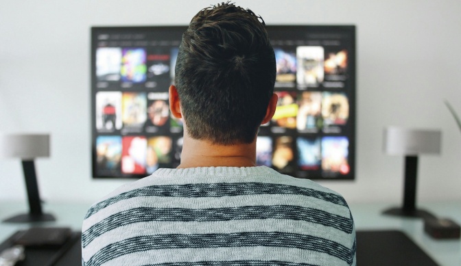Ein Mann sitzt vor einem Fernseher, auf dem verschiedene Filme in einem Auswahlmenü zu sehen sind.