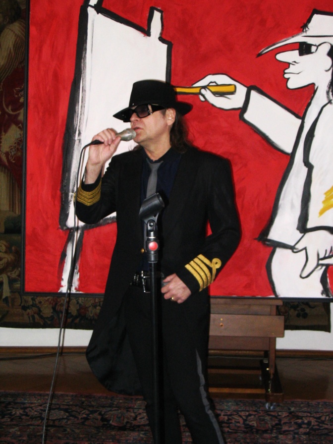 Udo Lindenberg steht vor einer bemalten Wand und singt in ein Handmikro. Er trägt einen Hut mit breiter Krempe und Gehrock.