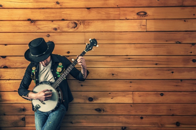 Ein Musiker mit Hut lehnt an einer Holzwand und spelt Banjo