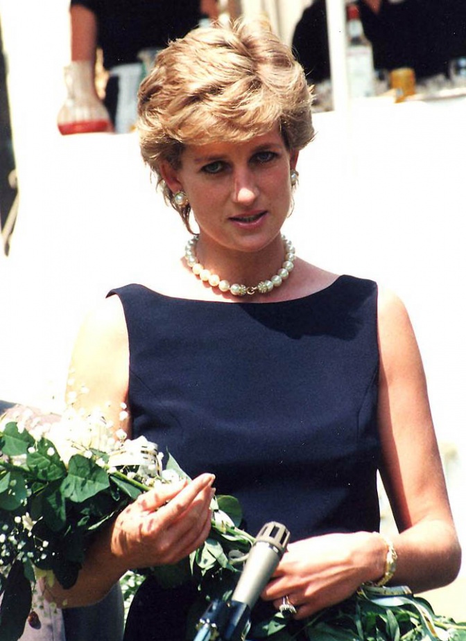 Prinzessin Diana mit kurzen, gewellten blonden Haaren. Sie trägt ein Etuikleid und Perlenkette und hält einen Blumenstrauß in der Hand.