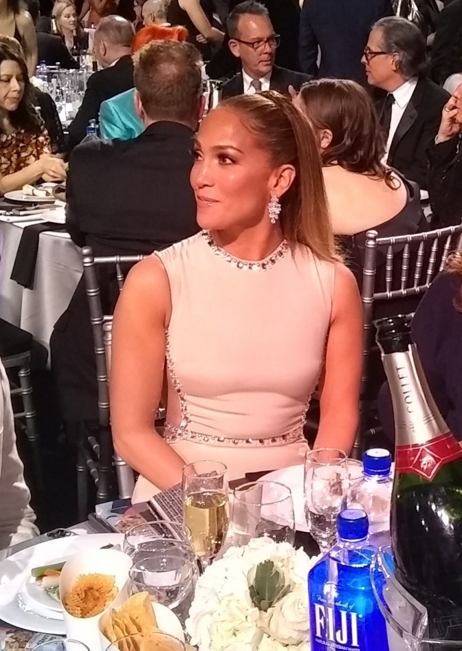 Jennifer Lopez sitzt in eleganter Kleidung vor einem Tisch mit Essen, im Hintergrund eine Menschenmenge