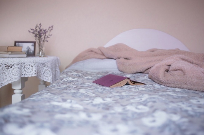 Ein aufgeschlagenes Buch und eine kuschlige Decke liegen auf einem Bett. Daneben steht ein Tischchen mit Spitzendecke und einem Blumenstrauß.