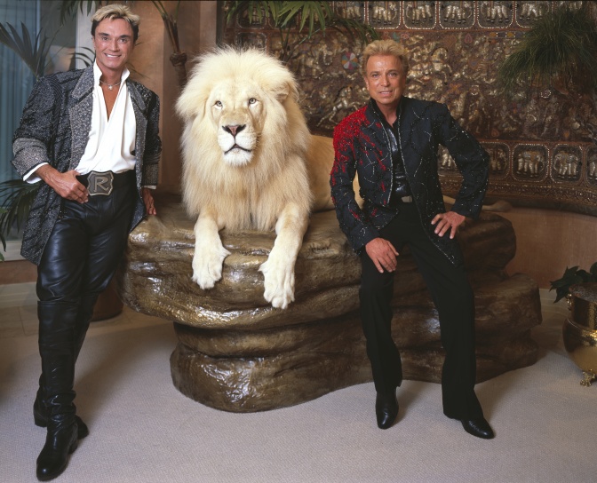 Siegfried und Roy stehen lächelnd neben einem weißen Löwen, der auf einem Podest zwischen ihnen liegt.