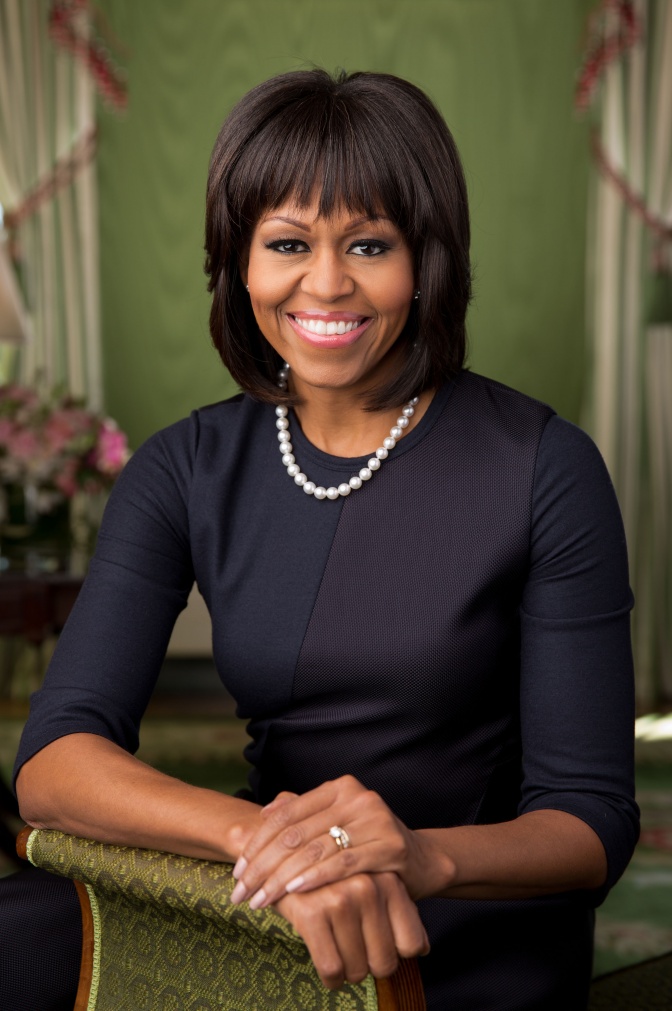 Michelle Obama mit kinnlangen, schwarzen Haaren und Pony. Sie trägt ein dunkelblaues Etuikleid und Perlenkette.