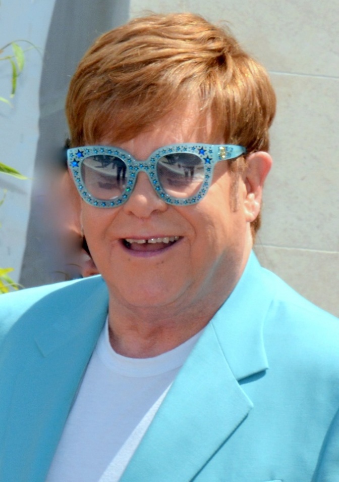 Elton John mit Föhnfrisur, in türkisem Sakko mit türkiser Brille.