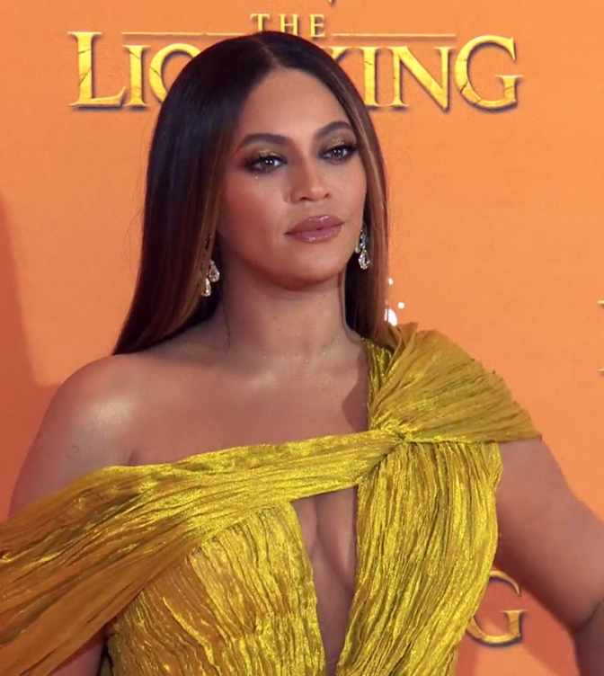Beyoncé mit langen, glatten, braunen Haaren. Sie trägt ein gerafftes, gelbes Abendkleid und steht vor einer Logowand mit dem Schriftzug Lion King.