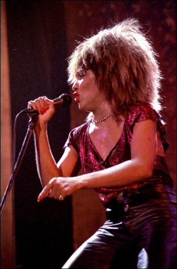Tina Turner mit Lockenperücke und im Pailettentop. Sie singt in ein Standmikro.
