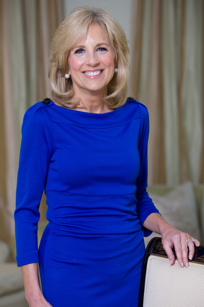 Jill Biden mit schulterlangen blonden Haaren. Sie lächelt und trägt ein schmales, leuchtend blaues Kleid.