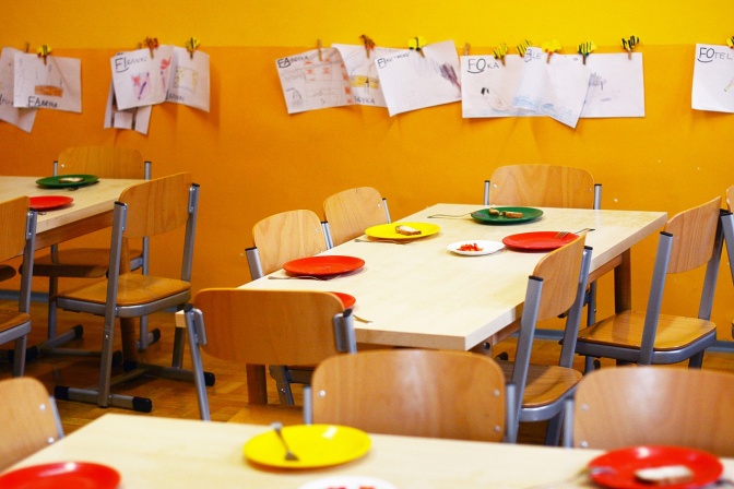 Kinderstühle und Tische in einer Kita. An der Wand hängt eine Leine mit Kinderzeichnungen.