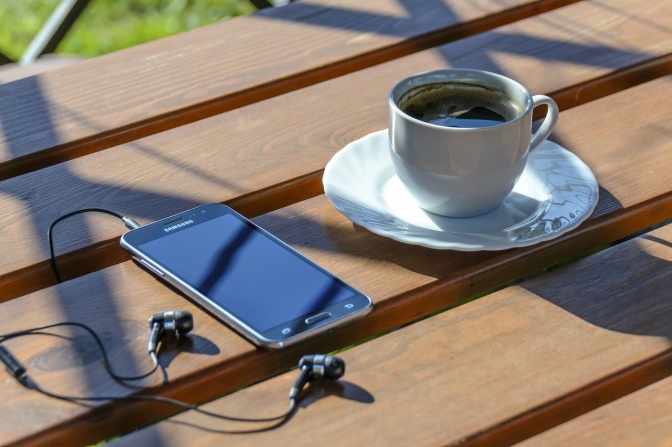 Ein Handy mit Kopfhörern liegt im Freien neben einer Tasse Kaffee.