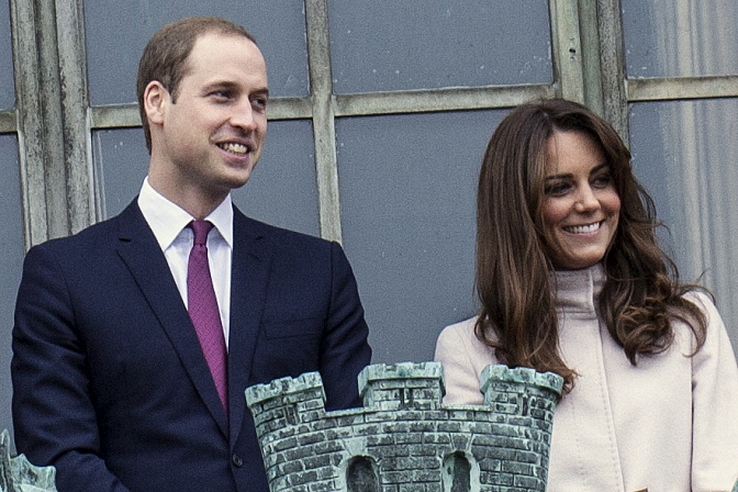 William und Kate stehen elegant gekleidet auf einem Balkon und lächeln. Kates Haare wehen im Wind.