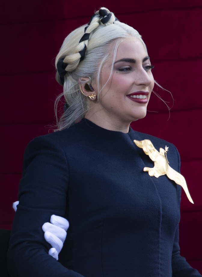 Lady Gaga mit weißblonden, geflochtenen Haaren. Sie lächelt. Auf ihrem Pullover ist eine Friedenstaube.
