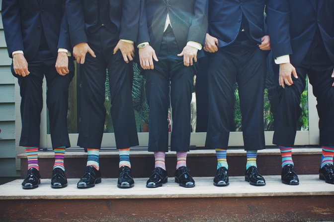 Eine Reihe von Männern steht nebeneinander. Sie tragen schwarze Anzüge und heben ihre Hosenbeine an. Sie tragen alle bunte Ringelsocken.