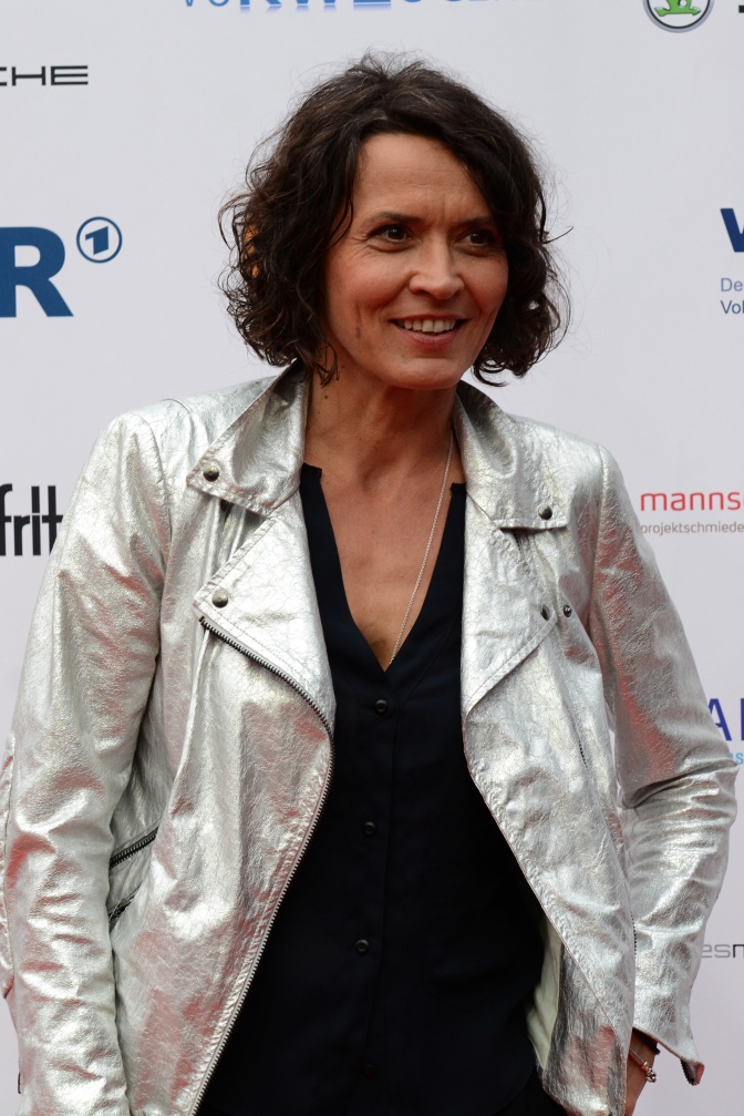 Ulrike Folkerts mit dunklen, gewellten kinnlangen Haaren in einer silbernen Lederjacke. Sie steht lächelnd vor einer Logowand.