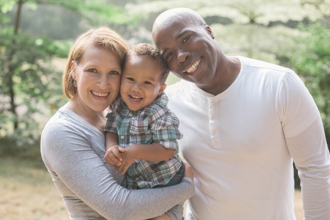 Ein Paar mit einem kleinen Kind im Arm. Alle 3 lächeln in die Kamera. Der Mann hat eine dunkle Hautfarbe, die Frau ist weiß.