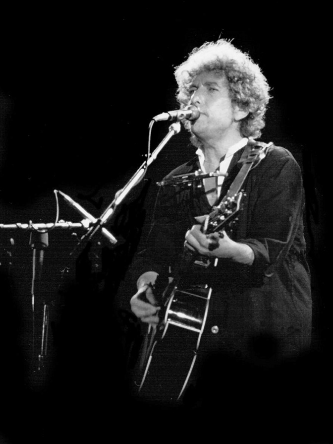Schwarz-weiß-Foto von Bob Dylan vor einem Mikrophon. Er singt und spielt Gitarre.