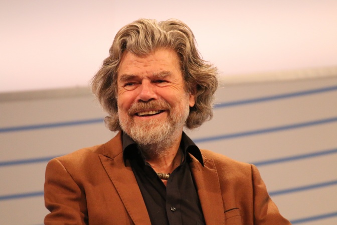 Reinhold Messner lächelnd in einem braunen Sakko. Seine Haare sind halblang und gewellt und werden grau.