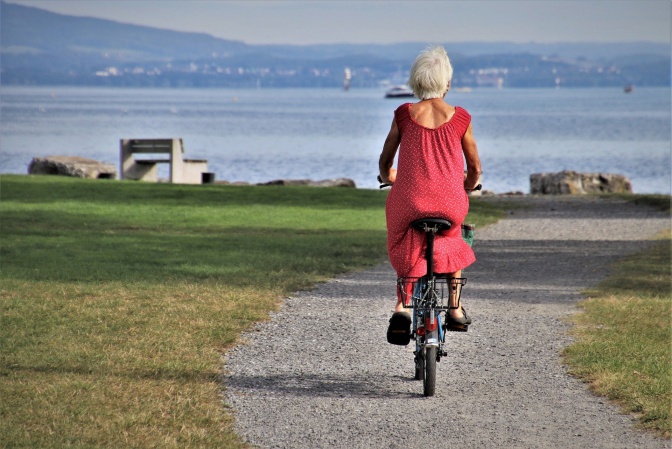 Eine ältere Frau mit kurzen weißen Haaren fährt auf dem Fahrrad. Im Hintergrund sieht man das Meer.