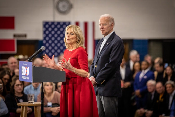 Jill und Joe Biden stehen nebeneinander an einem Rednerpult. Jill spricht, Joe hört zu. Im Hintergrund sitzen Menschen auf Stühlen um die beiden herum.
