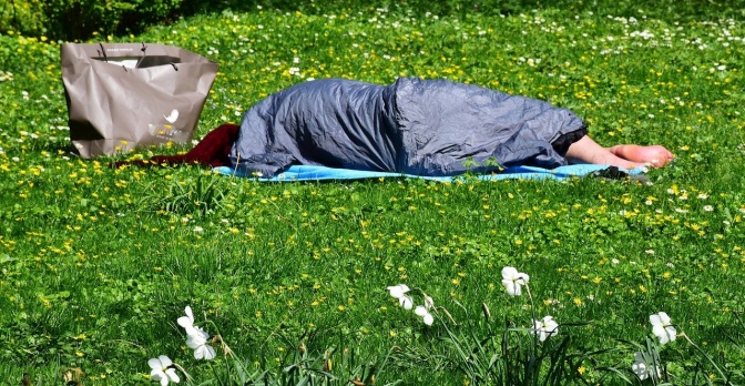 Eine Person liegt im Schlafsack auf einer Wiese, daneben stehen Plastiktüten.
