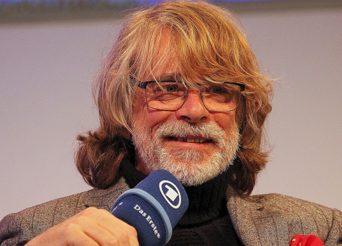 Helge Schneider mit schulterlangen, gewellten blonden Haaren, Brille und grauem Vollbart. Er spricht in ein Mikro mit dem Logo der ARD.