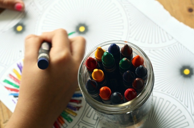 Eine Kinderhand hält einen Wachsstift in der Hand und malt ein Bild. Daneben stehen weitere Wachsstifte in einem Glas.