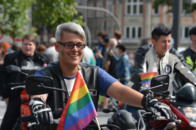 Mehrere Personen in Lederkluft auf Motorrädern. An einem Motorrad ist eine Regenbogenflagge befestigt.