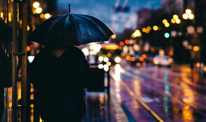 Ein Mann mir Regenschirm geht eine im Dunkeln beleuchtete Straße entlang.