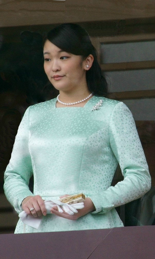 Eine japanische Frau mit Pferdeschwanz in einem eleganten türkisfarbenen, hochgeschlossenen Kleid. Sie trägt Perlenschmuck und hält Handschuhe und einen Fächer in der Hand.
