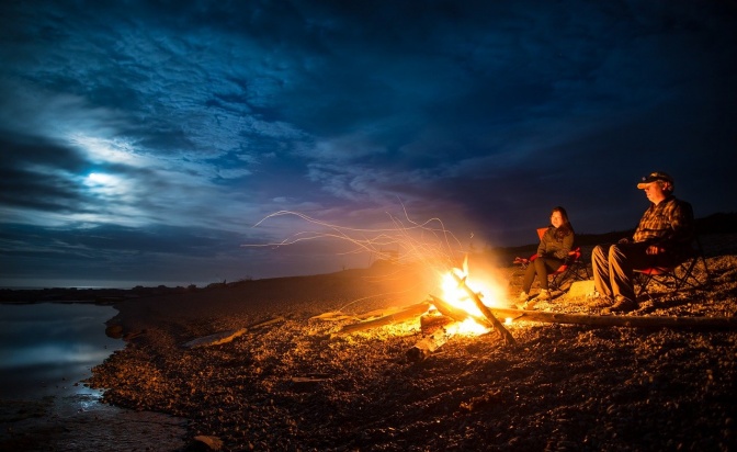 Zwei Personen sitzen an einem Lagerfeuer am Meer, über ihnen wolkiger, dunkler Himmel