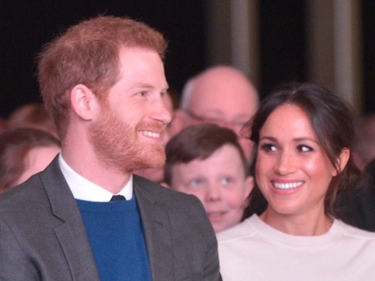 Prinz Harry und Herzogin Meghan sitzen nebeneinander und lächeln sich an.