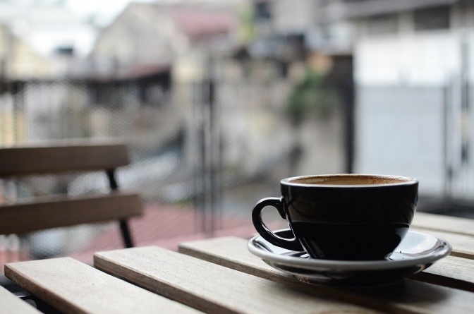 Eine Tasse Kaffee steht auf einem Tisch im Freien, im Hintergrund ein Stuhl.