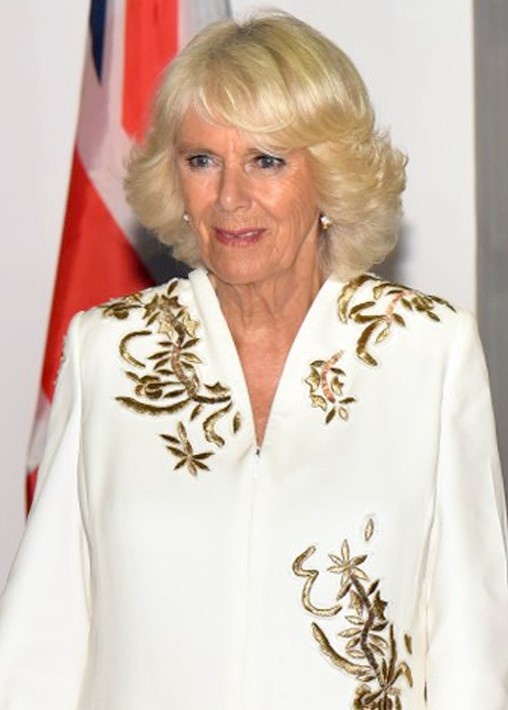 Herzogin Camilla in weißer Kleidung mit goldenen Ornamenten. Sie hat kinnlange blonde Haare und lächelt leicht.
