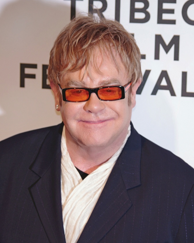 Elton John mit blonden Haaren und farbiger Sonnenbrille vor einer Logowand.
